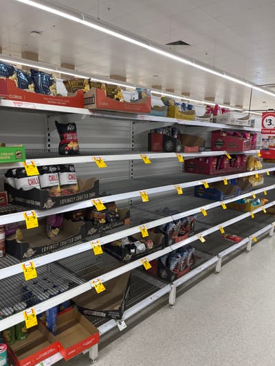 The image shows a grocery store shelf in Middle Park, Australia (latitude: -27.556, longitude: 152.923). The shelf is nearly empty, with only a few bags of chips remaining. The shelf is made of metal and has a grid pattern. There are yellow price tags on the shelf. The image is taken from a low angle, looking up at the shelf. The lighting is bright and the scene is clean. The empty shelves suggest that the store has been recently affected by a shortage or a surge in demand.  There are no people in the image.