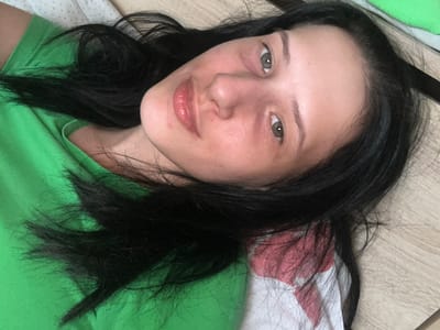 A close-up, overhead shot shows a young woman with dark hair lying on her back, her head tilted slightly to the right. She is wearing a bright green t-shirt. Her eyes are open and looking upwards, and her lips are slightly parted. The background appears to be a wooden surface with a patterned fabric beneath her head. The lighting is soft, creating a natural glow on her skin. The overall impression is one of relaxed repose, possibly taken in a bedroom or living area. The image does not contain any elements that indicate its location in Perushtitsa, Bulgaria.