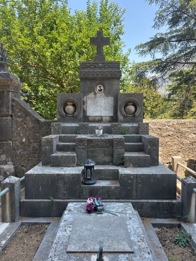The image depicts a large, tiered stone mausoleum situated in a cemetery in Škaljari, Montenegro. The mausoleum is constructed of grey stone and features multiple levels, ornate carvings, and decorative urns. At the top, a cross stands above a carved frieze and a small framed portrait. In front of the mausoleum, a lower stone slab serves as a tomb, with a small arrangement of purple and white flowers placed on it. A black lantern sits on a step of the mausoleum. The mausoleum is surrounded by lush green trees and vegetation, with a clear blue sky visible above. The overall impression is one of a well-maintained, albeit aged, monument within a serene cemetery setting.