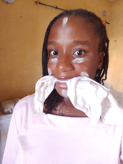 The image shows a close-up of a young Black woman, likely a teenager, with dark braided hair. She is wearing a light pink, square-necked top and a silver chain necklace. White cream or makeup is applied in streaks on her forehead, under her eyes, and on her cheeks, and a white cloth is held in her mouth, obscuring her lips. She is looking directly at the camera. The background is a textured, yellowish-orange wall, common in residential areas in Umuahia, Nigeria. The lighting suggests an indoor setting, possibly a bedroom or living area, with a hint of a bed and pillow visible in the blurred background on the left. The overall scene appears to be a candid, informal moment captured by a self-taken photograph.