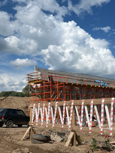 This image depicts a construction site in Kabanikha, Russia, featuring the ongoing construction of an elevated structure, likely a bridge or overpass. The foreground is dominated by a temporary barrier constructed from wooden beams and marked with alternating red and white stripes for visibility. Two old tires are stacked on the ground near the barrier. To the left, a black, early 2000s model Lada Niva is parked on the dirt ground.

In the mid-ground, the main construction activity is evident. Orange scaffolding forms a complex structure supporting a concrete deck, which is currently under construction. The concrete slab has rebar protrusions along its edge. Several support columns are visible beneath the deck, integrated with the scaffolding. A blue tarp is draped over a section of the deck. Behind the scaffolding, a dense line of green trees forms the background.

The upper portion of the image showcases a bright blue sky filled with large, fluffy white cumulus clouds, indicating fair weather. The overall scene conveys a sense of an active construction project under a clear sky.