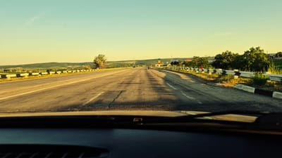 The image shows a road leading into the distance, with a clear, pale blue sky overhead. The sun is low in the sky, casting long shadows across the asphalt, suggesting it is either early morning or late afternoon. The road appears to be a highway, with multiple lanes and a guardrail on the right side. The landscape flanking the road is rolling hills covered in greenery, with scattered trees.
In the distance, there are signs and structures indicating a settlement. One sign is visible with the text "RAZENI" in white lettering on a blue background, confirming the location context as Razeni, Moldova. There are a few vehicles on the road, including cars and a truck, all moving in the same direction. The overall mood is one of travel and journey, with the warm, soft light contributing to a peaceful or serene atmosphere. The road surface shows signs of wear and tear, with cracks and patches visible. The foreground shows the dashboard of a vehicle, indicating the image was taken from the driver's perspective.