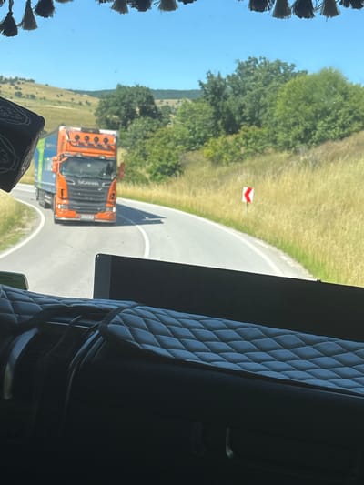 The image is taken from the interior of a vehicle, looking out through the windshield. In the foreground, the quilted dashboard and a portion of the driver's seat are visible. Outside, a curved asphalt road winds through a grassy, hilly landscape under a clear blue sky. On the road, an orange Scania truck is traveling away from the viewer. To the right of the road, a sharp curve warning sign is posted. The scene appears to be a rural road in the vicinity of Mladinovo, Bulgaria, given the landscape and the presence of a truck commonly used for long-haul transport. There are no people visible outside the vehicles.