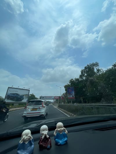 The image shows a dashboard inside a vehicle looking out onto a highway in Ahmedabad, India. In the foreground, three small monk figurines are positioned on the dashboard. The highway is visible with several vehicles, including a white Suzuki Ertiga with license plate GJ 01 T 4971, and a motorcycle on the left. Billboards are present along the roadside, one advertising a Maruti Suzuki Vitara and another for CP PLUS CCTV. The sky is partly cloudy with a light blue hue, and trees line the right side of the highway. The road has yellow and black striped markings on the shoulder.