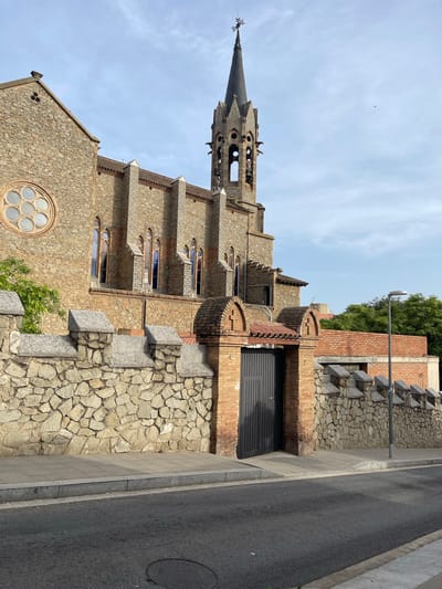 The image shows the exterior of a stone church building with a tall, pointed bell tower. The church has several gothic-style arched windows and a circular rose window on the left side. A stone wall with crenellated battlements runs along the front of the property, separating it from a paved sidewalk and an asphalt street. A dark gray metal gate is set within a brick section of the wall. Green trees are visible behind the wall and to the right of the church. The sky is partly cloudy. The building is identified as the Església de Sant Francesc d'Assís in Santa Coloma de Gramenet, Spain.