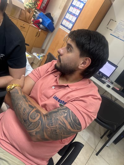A man with dark hair and a beard wears a pink Lacoste polo shirt. His left arm is covered in a detailed tattoo featuring architectural elements and an elephant design. He's wearing a gold watch on that wrist. Another person wearing a black shirt is partially visible in the lower left corner of the frame. The background includes office furniture like cabinets and chairs. A computer monitor is visible on a desk.