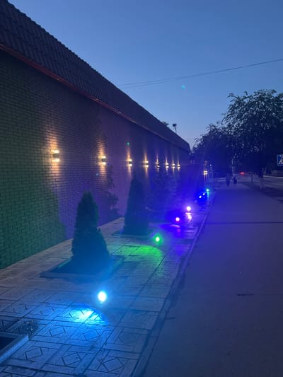This is a night-time photograph of a brick wall with modern outdoor lights illuminating it, located on a sidewalk in Temirtau, Kazakhstan. The wall, constructed of dark grey bricks, features several horizontally mounted light fixtures that cast a warm, yellow glow. Along the base of the wall, a row of small evergreen trees in rectangular planters are visible. The sidewalk, paved with decorative tiles, is lit by a series of colorful LED ground lights in shades of blue and green. A paved road runs parallel to the sidewalk. In the background, silhouetted trees and a few distant figures walking are visible under a dark blue twilight sky. A street sign is also present in the background. The overall atmosphere is tranquil and illuminated by artificial light.
