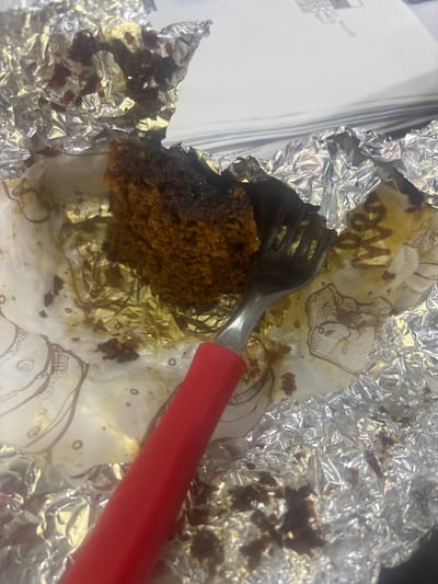 The image shows a close-up view of a partially eaten dark brown cake or muffin, presented in a white paper liner and wrapped in crumpled silver aluminum foil. A silver fork with a distinctive red handle is inserted into the baked good, suggesting it is actively being consumed or has just been eaten. The paper liner is stained with crumbs and oil from the cake and features faint brown illustrations and stylized letters, possibly "Me" or "W".
To the left of the food item, a stack of white papers or documents is visible, with some faint, illegible text and numbers. The word "DAVID" can be vaguely discerned on one of these papers. The overall setting appears to be an indoor environment, possibly a desk or table. No people are visible, and no specific time of day or weather conditions can be determined from the image. The scene captures a casual moment of a snack. While the context provides Buenavista, Mexico, there are no specific visual cues within the image itself to confirm this location.