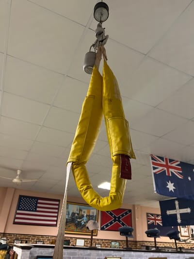 A yellow aircraft arresting gear pendant is suspended from the ceiling. Behind the gear are various flags including the United States, Australian, Confederate, and British flags, along with framed artwork and vintage hats. The room has a drop ceiling and a ceiling fan.