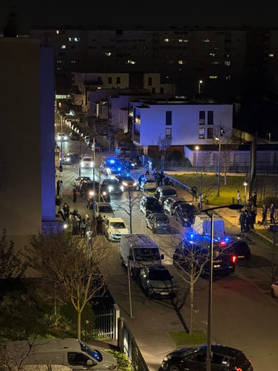 An elevated night-time view depicts a residential street in Bron, France. The street is lined with numerous parked civilian vehicles, including a white panel van in the foreground. At least four police vehicles display active blue flashing lights, one of which is a dark-colored van marked "POLICE NATIONALE" on its rear. One ambulance or emergency medical vehicle with blue flashing lights is also present. Approximately 20-30 individuals, primarily young adults, are gathered on the left sidewalk and partially on the roadway. On the right side of the street, multiple police officers in dark uniforms are observed interacting with smaller groups of individuals near the emergency vehicles. Modern residential buildings with illuminated windows line both sides of the street and form the backdrop under a dark sky. Streetlights provide general illumination, augmented by the emergency vehicle lights.