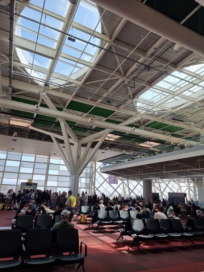 The image shows an indoor airport waiting area. The high ceiling features a complex network of white support beams and large windows that let in natural light. There is a digital display board mounted on the wall. Rows of black plastic chairs are arranged on a red carpet. Many people are sitting in the chairs, likely waiting for their flights. The location is not evident from the image. Therefore, determining that location in the city of Mora, Northern Cyprus / Cyprus is impossible.
