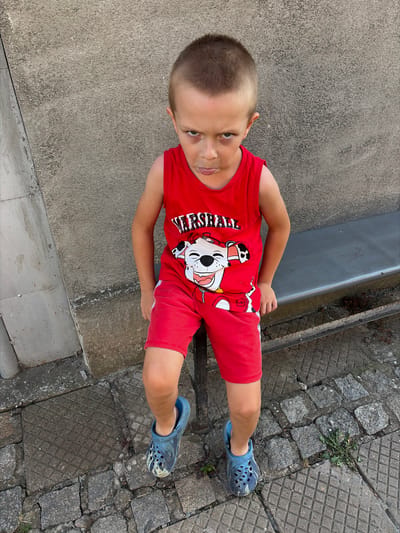A young boy, appearing to be around 5-7 years old, is sitting on a metal bench outdoors. He has short, light brown hair and is wearing a red tank top with a graphic of a dog on it and red shorts. He is also wearing blue Crocs. The boy is looking directly at the camera with a stern expression, sticking his tongue out slightly. The background consists of a textured concrete wall and a cobblestone ground. The scene appears to be in an urban or semi-urban setting. The image was taken in Dolni Dabnik, Bulgaria.