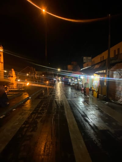 This nighttime image depicts a wet street scene in Kauman, Indonesia, observed from a low-angle perspective looking down a pedestrian street or narrow road. The ground, paved with alternating dark and lighter rectangular sections, is visibly wet, reflecting numerous light sources.

On the left side, in the mid-ground, a prominent golden-yellow structure with an ornate domed top, resembling a clock tower, stands tall. Below it, a low metal fence is visible, behind which a dark-clad individual is partially visible, kneeling or crouching. Further back, faint architectural elements and distant lights are apparent.

The right side features a series of brightly lit vendor stalls or small shops, sheltered by awnings. These stalls display various packaged goods. One stall is illuminated by a prominent light and has a sign above it reading "PUSAT REBANA." Behind the stalls, a multi-story building with yellow or beige walls is present. Utility poles with wires traverse the upper portion of the scene.

The center of the image is dominated by the wet street, which shows distinct light and dark striped patterns. Reflections from overhead streetlights and distant vehicle lights create elongated streaks across the wet surface. Several vehicles with illuminated headlights and taillights are visible in the distance, indicating traffic. The sky is dark, providing no natural light. A prominent overhead streetlight in the upper center emits a strong orange light, causing a significant horizontal lens flare across the image.