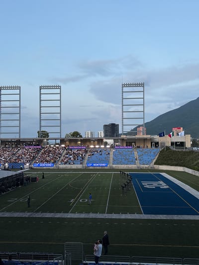 This is a wide, eye-level shot of a soccer stadium in Monterrey, Mexico, likely taken during an evening event. The sky is a pale blue with some scattered clouds. In the foreground, the green turf of the soccer field is visible, marked with white lines and numbers indicating yardage. Several spotlights on stands are positioned on the field. To the right, a blue end zone with a large white logo, possibly for a team or university, is prominent. In the midground, bleacher seating is filled with a large crowd of spectators dressed in white. Signs reading "Hector Martin Perez Borjas" and "LCP-MONTERREY" are visible above the seating sections. Three tall, skeletal metal structures with rows of lights at the top are positioned behind the bleachers, reaching towards the sky. In the background, several buildings of varying heights, including a modern skyscraper, are visible, and a large, hazy mountain dominates the right side of the frame. Two flags, one of Mexico and another blue flag with a white symbol, fly above a building on the right. Several people are scattered across the field, including two men in suits and a man holding a child in the lower right section of the frame. The overall impression is of a bustling sporting event.