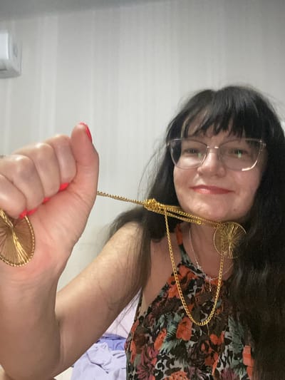 A person is shown in a close-up, overhead shot. The subject has long dark hair, glasses, and is wearing a floral-patterned top. They are also adorned with prominent gold jewelry, including large hoop earrings and a layered necklace. The person is holding a gold ring in their right hand, which is close to the camera, obscuring part of their face and the lower half of the image. Their fingernails are painted bright red. The background is a plain, light-colored surface, possibly a wall or ceiling, with some indistinct lines that could suggest a textured finish or wallpaper. The lighting appears to be indoor, with a soft, even illumination. No specific time of day or weather conditions can be determined from the image. The overall mood is somewhat casual, given the selfie-like perspective and the focus on personal adornment. There is no visible text in the image. The location context of Novi Petrivtsi, Ukraine, does not provide any specific visual cues within the image itself.
