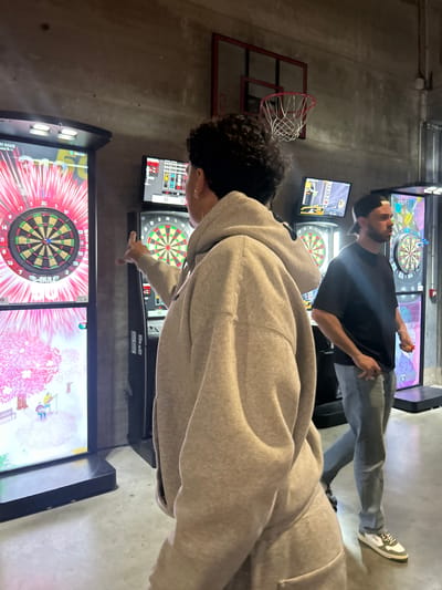 This media file captures an indoor scene in Villeneuve-d'Ascq, France, featuring two young men engaged in playing electronic darts. The image is taken from a slightly elevated perspective, looking across the room.
In the foreground, a man with curly dark hair, wearing a light grey hooded sweatshirt, is positioned to the left. He is seen pointing towards one of the electronic dartboards, seemingly observing the game or indicating something on the screen. Further back and to the right, another young man, dressed in a black t-shirt and grey jeans, stands facing a separate dartboard. He appears to be in the process of throwing or aiming a dart, with his body turned and arm extended.
The setting is a recreational space, likely an arcade or a bar equipped with various games, indicated by the multiple electronic dartboards and a miniature basketball hoop with a red backboard visible on the left. The room has an industrial aesthetic, characterized by concrete walls. Each dartboard station features screens displaying colorful game interfaces. Visible text on these screens includes "01 GAME," "UND 5/15" (likely "ROUND 5/15"), and "ONLINE." Other game-related numbers like "15-BUL 2," "57," and "73" are also present, along with the traditional numbers on the dartboard itself (e.g., 20, 14, 11, 8, 16, 15, 10). The atmosphere appears casual, with both individuals focused on their game.