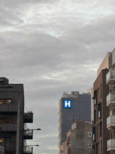 The image depicts a view of the sky above Aubervilliers, France, framed by buildings. In the foreground, the upper portion of a grey building with horizontal lines is visible, punctuated by windows and rooftop structures, including streetlights and what appears to be mechanical equipment. Above and slightly to the right, a taller, grey building has a large blue square with a white "H" on it, likely indicating a hospital or medical facility. Above this, the upper portions of several other buildings of varying heights and architectural styles are visible, some with balconies and others with what appears to be brick facade. The sky is overcast with heavy cloud cover, giving the scene a subdued, neutral color palette.