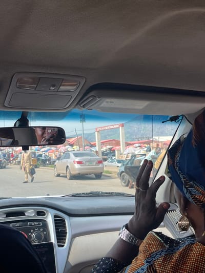 The image is a view from the interior of a car, looking forward through the windshield. The car appears to be stopped or moving slowly in traffic. Through the windshield, a busy street scene is visible, with a sign that reads "DUTSEN ALHAJI TRADITIONAL MARKET." Numerous stalls with colorful umbrellas are set up, and various people and vehicles, including motorcycles and cars, are present. In the foreground, a woman's hand, wearing a watch and rings, is visible on the right side of the frame. A rearview mirror is attached to the windshield, reflecting a person holding a phone and taking a selfie. A man in a tan traditional outfit is walking across the street in the middle ground. The sky is overcast, and the overall lighting suggests daytime.