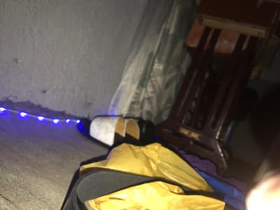 The image depicts an indoor scene in Kirana, Nigeria, under dim illumination. A string of active blue LED lights extends horizontally along the base of a light grey, textured wall. To the right of the lights, two partially rolled or folded white and black objects, possibly footwear, are situated. In the immediate foreground, a crumpled yellow plastic bag rests beside a dark object with a visible wide strap. In the background, a dark reddish-brown wooden or metal furniture frame, resembling a sewing machine stand, is partially visible against a patterned fabric backdrop. No individuals are present in the scene.
