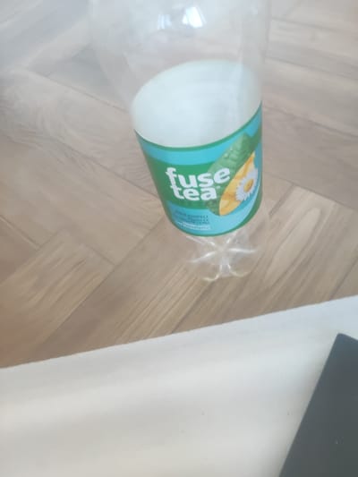 The image shows a close-up, overhead view of a clear plastic beverage bottle on a wooden floor. The bottle has a green and blue label that reads "fuse tea" and features an illustration of mango and chamomile. The floor appears to be made of light-colored wood planks laid in a herringbone pattern. A white, textured surface is visible in the foreground, and a dark rectangular object, possibly a book or electronic device, is partially visible at the bottom right edge of the frame. The lighting suggests an indoor setting. The image does not provide any specific clues to determine its location in the city of Bukhara, Uzbekistan.