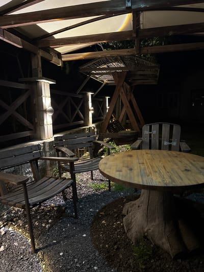 The image depicts a rustic outdoor seating area at night, situated in the coastal town of Tyulenovo, Bulgaria. A wooden table with a rough-hewn tree trunk base is centrally positioned, surrounded by several wooden chairs and benches. The seating is arranged on a gravel and dirt ground beneath a wooden and fabric canopy, which is illuminated by artificial lighting. In the background, a wooden swing set is visible, also appearing to be made of rustic materials. A wooden fence with crisscross patterns runs along the side of the area, with wall-mounted lights attached to stone pillars. The overall scene suggests an outdoor dining or relaxation space, likely part of a guesthouse or rural accommodation, given the natural and unrefined aesthetic. The lighting creates shadows, enhancing the nighttime ambiance. There are no people visible in the image.