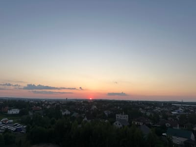 This is an aerial, high-angle, panoramic photograph of a suburban landscape during a sunset, likely taken from a considerable height. The sky exhibits a gradient of colors, transitioning from a clear pale blue at the top to soft orange and pink hues near the horizon. A distinct, bright red sun is visible just above the horizon, partially obscured by a thin band of clouds.
The foreground is dominated by the dark silhouettes of numerous deciduous trees, creating a dense, green canopy that obscures the ground below. Beyond the trees, a town or village unfolds, characterized by a dense collection of residential buildings with varied roof colors, including red, grey, and white. The architecture appears to be primarily houses with pitched roofs, suggesting a typical residential area.
In the mid-ground, a street is visible, lined with parked cars, including several passenger vehicles and a few larger vehicles like vans and a truck. A water tower or a similar tall structure is identifiable on the left side of the horizon. In the far distance, the landscape appears flatter with some scattered structures and the faint outline of more distant vegetation or terrain. The overall impression is one of a peaceful, quiet evening in a rural or semi-rural setting, characteristic of the Шчомысліцкі сельскі Савет, Belarus. There are no people visible in the image.