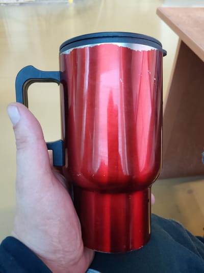This is a close-up, eye-level shot of a hand holding a shiny red travel mug. The mug has a black lid and a black handle attached to its side. The mug appears to be metallic and has vertical ridges or indentations running down its body. The hand holding the mug is fair-skinned and positioned in the lower left quadrant of the frame. The background is blurred, but hints at a yellow-toned surface, possibly a table or floor, and a brown, wooden object on the right, which could be furniture. The lighting suggests an indoor setting. There is no visible text in the image. The overall impression is of a casual, everyday moment.