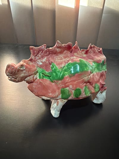 An artistic, ceramic sculpture of a pig-like creature with a long snout and sharp dorsal spines is positioned on a dark, flat surface. The creature is primarily colored in pink and red, with prominent green stripes and splotches across its body. Its four stubby legs are white, and its eyes are small and round. The background consists of vertical, textured panels that are lit from above, creating a soft, diffused light. The image is taken from a side angle, with the creature's head facing left. The overall impression is of a whimsical, handmade art piece. This sculpture is located in Bissendorf, Germany.