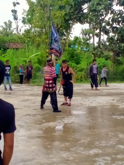 The image depicts a group of men in a concrete outdoor space, possibly a schoolyard or a community gathering area. The men are standing in a circle, facing each other, and there is a flagpole in the background. There is a man in the center holding a whip or stick in his hand, with another man standing nearby. The rest of the group are standing around the edges of the circle, seemingly observing. There is a sign with the words "Tarate" on it, possibly related to a local martial arts club. The ground is wet, and the vegetation surrounding the concrete area appears to be green and lush. The location of the image, based on the coordinates provided, is in the Kecamatan Kedawung area of Indonesia.
