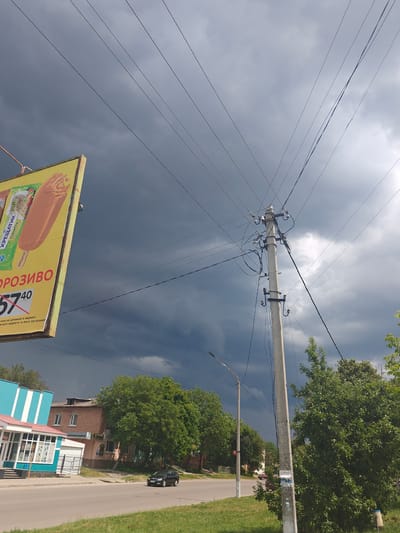 The image depicts a street scene in Hadiach, Ukraine, under a dark, cloudy sky suggestive of impending rain. A prominent billboard advertising ice cream, with the text "ХРЕЩАТИК ПЛОМБІР" and a price of "57 40," is positioned on the left side of the frame. The billboard is yellow and features an image of a chocolate-covered ice cream bar. Overhead, a complex network of power lines and a concrete utility pole are visible. In the background, a street runs alongside a sidewalk, with several buildings, including a blue and white structure with an awning, and a brick building. Green trees line the street and sidewalk, with a black car driving on the road. The overall atmosphere is overcast and possibly humid.