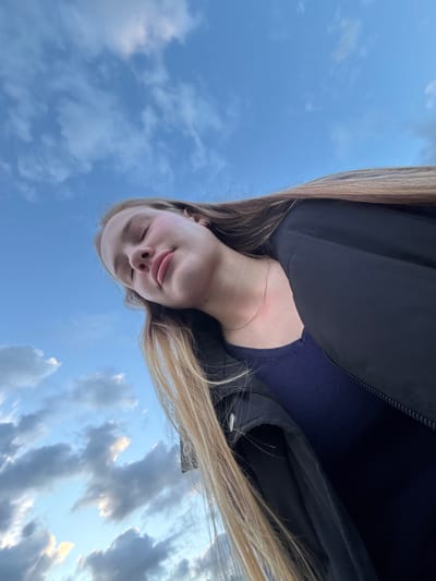 A low-angle, medium shot captures a young person with their eyes closed, tilted slightly upwards against a blue sky with scattered clouds. Their face is turned to the side, with blonde hair falling across their shoulder. They are wearing a navy blue shirt and a black jacket. A thin silver necklace is visible around their neck. The location is implied to be Solnechnoye, Russia, suggested by a contextual element not present in the visual data itself. The overall mood is serene and contemplative, with the focus on the person's peaceful expression and the expansive sky.