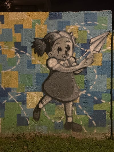 This media file captures street art on a textured wall, likely concrete blocks, in Engomi̇ Lefkoşa, Cyprus. The central subject is a stylized, cartoon-like figure, resembling a child or girl, painted in shades of gray and black with white highlights. The figure has large, round eyes from which black liquid appears to be dripping, conveying an impression of sadness or tears. It is depicted holding an inverted, umbrella-like object above its head.

The background of the graffiti features a vibrant checkerboard pattern composed of varying shades of blue, green, and yellow squares. White dashed lines are overlaid on this patterned background, suggesting movement or a dotted path. To the right side of the painted wall, natural green grass is visible, indicating an outdoor setting. The lighting in the image appears to be dim, possibly suggesting it was taken at dusk or night, with some artificial light sources illuminating the artwork. No explicit text is visible within the image.