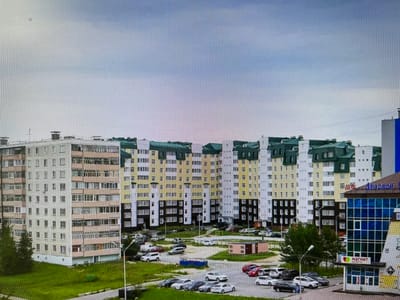 This is a wide, eye-level, outdoor shot taken on an overcast day, showcasing a street view of a residential area in Pyt'-Yakh, Russia. The central focus is a row of multi-story apartment buildings, characterized by their beige, yellow, and white facades, with dark-colored window frames and green accents on the upper levels. These buildings exhibit a consistent architectural style, with numerous balconies and windows.

To the right, a modern, multi-story commercial building with a blue glass facade stands out. A sign on this building, in Cyrillic script, reads "Магнит," indicating a retail store. A smaller, darker building with a peaked roof is situated behind it.

In the foreground, a grassy area slopes down towards a paved road or parking lot. Several cars are parked in the lot, including white SUVs, sedans in various colors (black, silver, red), and a white van. Some of the cars appear to be in motion, suggesting ongoing activity. A lamppost with a single light fixture stands prominently in the middle ground. Further to the left, more trees and greenery are visible, with a large, older-style apartment building partially obscured by them. The sky is predominantly cloudy with hints of blue. The overall impression is one of a developed urban residential and commercial landscape.