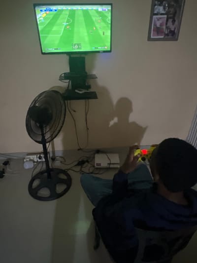 The image captures an indoor scene, likely a casual living space such as a living room or bedroom, with the perspective tilted. A person is the central subject, seen from a slightly elevated and side angle, engrossed in playing a video game. They are wearing a dark hoodie over a lighter shirt and possibly blue jeans, with a dark backpack slung over their shoulder. The person is crouched or kneeling, holding a brightly colored yellow and black video game controller that emits a red light. A distinct shadow of the person is cast on the wall behind them.

To the left of the person, a flat-screen television is displaying a football (soccer) video game with a green pitch and player icons. Below the TV, a large black standing floor fan is visible. Behind the fan and beneath the television, a wall-mounted shelf holds several electronic devices, possibly a gaming console or router, surrounded by a tangle of cables and wires. A power strip with multiple plugs is visible on the floor, connecting various electronics. Above the television, a rectangular picture frame containing several smaller photographs is mounted on the wall.

The lighting in the room is dim, suggesting the time of day is either evening or night, with the glow from the television screen and the game controller providing some illumination. The scene depicts a moment of focused entertainment. No specific text is clearly legible from the visible elements like the picture frame or the game interface.
