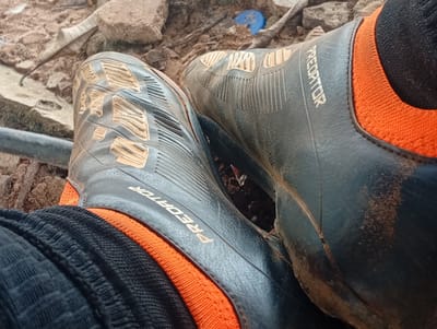 The image shows a close-up, overhead view of two feet wearing dark athletic shoes with orange ankle cuffs and black socks. The shoes have the word "PREDATOR" embossed on the side in gold lettering. The soles of the shoes have a textured pattern in a gold color. The shoes appear to be dirty, with some brown soil visible on the fabric and the sides of the shoes. The background consists of dry soil, rocks, and some debris, suggesting an outdoor, possibly unpaved, environment. There is no clear indication of people or specific actions occurring, other than the feet being present in the shoes. The image does not provide enough visual information to determine a specific location within Ukukwa, Nigeria, or any city.