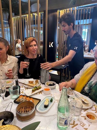 The image shows an interior scene, likely at a restaurant in Torreforta, Spain. A man wearing a black t-shirt is pouring a grey bottle of liquid into a glass held by a woman with red hair, who is seated at the table. Other people are visible at the table, including a woman in a white shirt with elaborately painted fingernails, and a woman wearing a yellow and floral patterned garment. The table is covered in sushi dishes and other food items. A partially consumed plate of food with utensils is visible at the top of the frame, and several water bottles and glasses are also visible on the table. In the background, there are decorative gold-colored bars creating a screen-like effect. The image is slightly tilted.