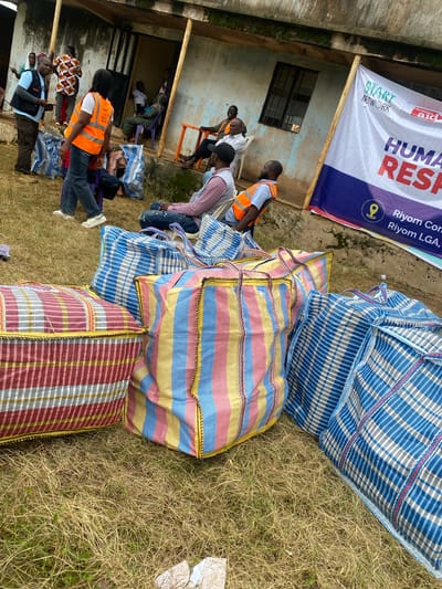 This image depicts a scene in Riyom, Nigeria, likely related to a humanitarian aid distribution. Several people are gathered around a building, some sitting and others standing. Many are wearing high-visibility vests, suggesting they are volunteers or aid workers. The event appears to be taking place outdoors, with dry grass and what looks like dirt or gravel on the ground.

Dominating the foreground and right side of the image are large, colorful woven bags, some with blue and white checkered patterns and others with stripes of red, yellow, blue, and white. These bags are likely filled with supplies for distribution.

In the background, a banner is prominently displayed, featuring text related to "START NETWORK" and "CHRISTIAN AID," indicating the organizations involved. The banner also has text reading "HUMAN RES[P]" which is likely part of "HUMANITARIAN RESPONSE". Further text on the banner mentions "Riyom Con" and "Riyom LGA," confirming the location. The presence of the banner, the volunteers in vests, and the aid bags strongly suggest a humanitarian aid distribution event.

The overall mood appears to be functional, with people engaged in activities related to the aid effort. The lighting suggests it is daytime, and the dry grass indicates it might be a dry season.