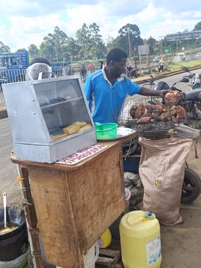 The image shows a street vendor selling food in Githurai, Kenya (latitude: -1.201, longitude: 36.92). The vendor, a man in a blue polo shirt, is standing behind a wooden food stall, facing right. He is preparing food, likely sweet potatoes, by grilling them on a small charcoal grill. The grill is situated on top of a small table in front of the stall. The food is arranged on a wire rack above the grill. Several sweet potatoes are on the rack, some already cooked and some still grilling. The vendor is using his left hand to pick up cooked sweet potatoes from the rack. The stall is equipped with a metal display case, possibly for showing other food items. The case is empty. A green plastic container, likely used for washing hands, is placed on the table. Behind the stall, there is a large bag of charcoal, which is next to a dark-colored plastic container. A yellow plastic container of liquid, likely water, sits in front of the stall. The vendor is wearing a white cap, and his hands are visible. The vendor is located in the middle of the image. The street environment is a busy intersection, with a road and sidewalks. Traffic can be seen in the background. The sky is cloudy, and the sun is shining. The vendor is preparing food for customers who are not visible in the image.