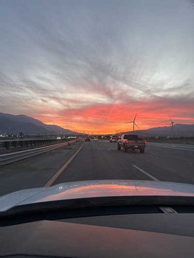 The image depicts a multi-lane highway in the United States, specifically in the vicinity of Desert Hot Springs. The view is from inside a vehicle, showing the hood of the car in the foreground. Several other vehicles are visible on the highway, including a truck in the adjacent lane. The sky is dominated by a vibrant sunset, with hues of orange and pink. In the distance, mountains are visible along with wind turbines, indicative of a wind farm. A roadside barrier is present on the left side of the highway.