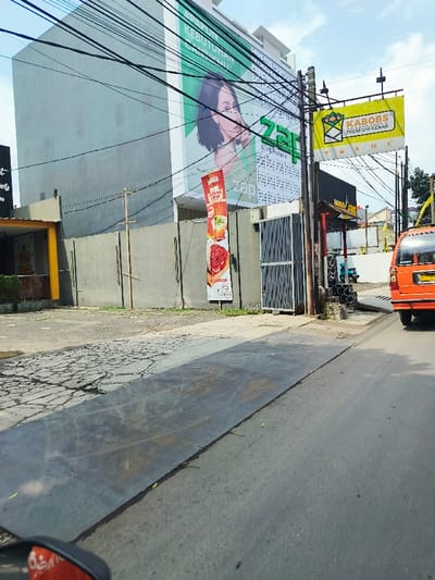 The image shows a street scene in Cimahi, Indonesia, taken at the intersection of Jalan Raya Cimahi and Jalan Setiabudi (latitude: -6.869, longitude: 107.533). The street is paved with asphalt and has a metal plate in the foreground. A gray building with a large billboard advertising a beauty product faces the street. The billboard depicts a young woman with long dark hair looking directly at the viewer. Another billboard advertising a restaurant called “Kabobs Premium Kebab” is mounted on a metal pole. A red and orange van is driving away from the viewer on the left side of the road. The van is parked near a closed metal gate with a white wooden sign above it. The building is surrounded by a chain link fence. Power lines run along the front of the building. There are no people visible in the image. The sky is clear and blue with white clouds.