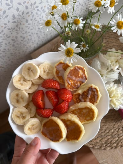 A hand is holding a white, scalloped-edge plate filled with a breakfast assortment. The plate contains sliced bananas, halved strawberries, and small, golden-brown fritters drizzled with a white sauce. In the background, a bouquet of white daisies with yellow centers is in a glass vase, and a textured burlap placemat is visible. The scene appears to be indoors, with a patterned wallpaper in the background. The location is not specified in the image.