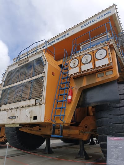 This is a medium-shot, eye-level, outdoor photograph of a massive yellow and white mining dump truck, specifically a BelAZ-75600. The truck is the primary subject, dominating the frame. It is stationary, likely on display or in a museum, supported by black stands. The truck's colossal tires are a prominent feature, with one clearly visible on the right side of the image, showcasing its immense size.

The truck has a bright orange cabin area with large, circular gauges and a blue ladder leading up to the driver's platform. A red fire extinguisher is mounted on the side of the ladder. The front of the truck features large, grilled vents. The sky above is partly cloudy and appears to be daylight, though the exact time of day is not clearly discernible.

Visible text on the truck includes "#AUTO_UGMK" and "#auto_ugmk" in blue lettering on the upper structure. There is also some Russian text on a sign placed near the front tire. This sign provides details about the "Карьерный самосвал БелАЗ-75600" (Mining dump truck BelAZ-75600), listing its specifications and indicating it was in production from 2005 to the present time. The location context provided is Verkhnyaya Pyshma, Russia. No people are visible in the image. The scene suggests a static display of industrial machinery.
