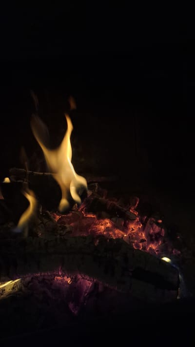 The image depicts a close-up view of a fire. The scene is dark, dominated by a black background. On the right side, there is a burning log with glowing embers and flames. The log appears to be partially charred. The flames are mainly yellowish-orange and appear blurred, suggesting movement. There are no people present in the image. The location is Profesor Ishirkovo, Bulgaria.