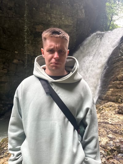 A close-up, medium shot shows a man standing outdoors in a rocky, shaded area with a waterfall in the background. The man is fair-skinned with short, light brown hair and a serious expression. He is wearing a light gray hooded sweatshirt with a black strap across his chest. The location is in Tsabal, Abkhazia, identified by the distinct geological features and waterfall in the background. The environment appears damp and rocky, indicative of a natural canyon or grotto near the waterfall. The photo is taken from a slightly low angle.