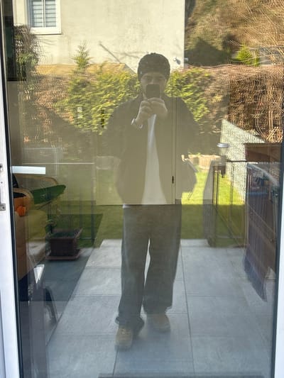 This image displays a full-length reflection of an adult male captured in a large glass patio door, located in a residential area of Hagen, Germany. The male subject, positioned centrally, is wearing a dark beanie, a dark collared jacket over a white t-shirt, dark trousers, and light-brown work boots. He is actively holding a smartphone with both hands at chest level, indicating the action of taking a selfie. A light-colored wristwatch is visible on his left wrist.

Through the glass, behind the reflection, the immediate outdoor environment consists of a light-gray tiled patio, transitioning into a manicured green lawn. A dark metal fence, topped with a solar-powered light fixture, runs along the perimeter of the lawn. Beyond the fence, a dense green hedge of coniferous trees is visible, interspersed with dormant deciduous trees, suggesting a late winter or early spring season. In the background, a light-colored building with a white-framed window featuring horizontal blinds is present, backed by a sparsely vegetated hillside. The sky appears clear and bright, indicating sunny conditions.

Reflected on the interior side of the glass, faint details of an indoor setting are discernible to the left, including a light-colored armchair and a potted plant in a brown terracotta pot. The indoor flooring beneath the reflection is also a light-gray tiled surface.