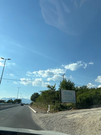 The media file displays a roadside scene under a clear blue sky dotted with white, puffy clouds, suggesting a sunny day. The perspective is from inside a vehicle, looking out towards a road that stretches into the distance, flanked by green trees and shrubs on the left and a gravelly verge on the right. In the distance, mountains are faintly visible, hinting at a rural or semi-rural mountainous region.
Several streetlights line the side of the road, and power lines run overhead. A sign is mounted on a pole to the left of the road, partially obscured by trees. The sign features a logo and text, which appears to read "SMART" and "project>design> (construction)". Below this, a phone number is visible: "+382 67 380 777".
Vehicles are traveling on the road, indicating it is a functional route. The overall impression is one of travel on a bright, possibly warm, day in a scenic area. There are no people clearly visible, but the context suggests a journey or transit. The time of day appears to be late morning or early afternoon, given the bright sunlight. The scene evokes a sense of movement and openness, typical of a road trip.