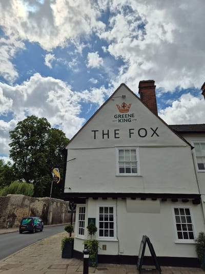 This outdoor image captures the facade of "The Fox" pub, a traditional British establishment, under a partly cloudy sky during the daytime. The building, painted in a light cream or white color, features multiple white-framed windows with small panes and dark trim. The name "THE FOX" is prominently displayed vertically on the side of the building in large black letters, accompanied by the "GREENE KING ESTº 1799" logo, which includes a stylized crown. A brick chimney is visible on the roof.

A hanging sign near the pub's entrance also features "THE FOX" and a stylized fox head in black and yellow. The pub is situated alongside a paved street, where two dark-colored vans are parked. Partial text like "VERSO" and "VEGA" along with green and blue graphics can be seen on the vans. A robust stone wall, suggesting historical architecture, runs parallel to the street opposite the pub. Green trees and foliage are abundant around the area, contributing to a natural setting. Potted plants add decorative touches to the pub's exterior.

The sky is a mix of bright blue and white, fluffy clouds, indicating fair weather. The scene is quiet, with no people visible or specific activities taking place, portraying a calm moment in what appears to be a historic village or town setting in West Suffolk, United Kingdom.