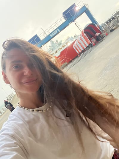 A young woman with long brown hair is taking a selfie outdoors, with a port or industrial setting in the background. The image is oriented sideways. She is wearing a white t-shirt and a necklace made of shells. Her expression appears relaxed and pleasant.
In the background, a large red and white structure, possibly a ship or part of a crane, is visible. A blue gantry crane with the text "RAMP 2" is also prominent in the scene. The sky is bright, suggesting it is daytime, and the weather appears to be fair. The overall setting seems to be a busy port area, given the industrial equipment. A small portion of another person is visible in the lower right corner, wearing a dark jacket with colorful graphics. The location context provided is Don Sak, Thailand.