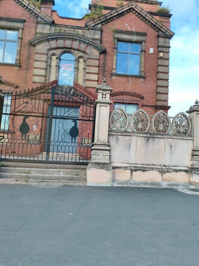 The image shows the exterior of a brick building in Sandwell, United Kingdom, with "SCHOOLS" written above a large arched window. A black metal gate with decorative elements and two silhouette figures of birds stands in front of the building. A light-colored stone wall with ornate decorative panels runs along the side of the building, and a paved road is visible in the foreground. The sky is blue with some clouds.