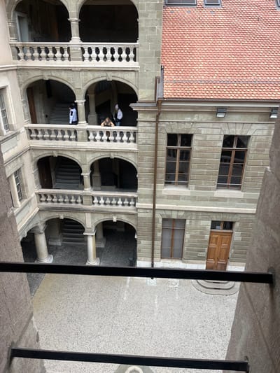 The image presents a view looking down into a large, ornate stone courtyard or atrium in Geneva, Switzerland, captured from a high vantage point behind dark metal railings. The architecture is grand and historic, featuring multi-story buildings constructed from light-colored, neatly cut stone blocks. These structures are characterized by a series of stately arched openings that form walkways or loggias on different levels, supported by robust columns and decorated with intricate balustrades.
Several individuals are visible on these upper levels. On a lower arched walkway, one person in a white jacket and dark attire is standing. Higher up, two other people are present on an open balcony: one with light-colored hair and a dark top, and another in a lighter-colored top, both seemingly observing the courtyard below or moving through the space.
To the left (when viewed upright), an adjacent building is topped with a distinctive red-tiled roof, characteristic of European architecture. The opposite facade features multiple rectangular windows with wooden frames. An unusual architectural detail is a large wooden double door integrated into a window opening on one of the upper floors. The ground level of the courtyard, partially obscured, appears to be paved with light-colored, textured material.
The lighting is diffuse daylight, suggesting an overcast day or perhaps morning/late afternoon, casting soft shadows that highlight the architectural depth. The overall atmosphere is one of quiet observation within a detailed and historically rich urban environment. There is no visible text in the image.
